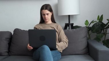 Focused young brunette female freelancer sitting on cozy sofa, working distantly on laptop, solving problems online, making difficult decision, looking in distance searching for inspiration at home