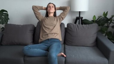 Relaxed beautiful young woman 20s put hands behind head resting at home leaned on sofa cushions. Modern apartment owner, air conditioner, climate control indoor, no stress, lazy weekends concept