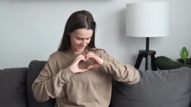 Sincere beautiful millennial young caucasian woman looking at camera, showing heart symbol, expressing love, feeling affection. Candid female volunteer demonstrating donation sign sitting on sofa