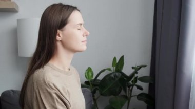 Calm young woman taking deep breath of fresh air meditating with eyes closed sitting on cozy sofa near window at home. Enjoy moment of peace, do mental relaxation exercises, feels no stress concept