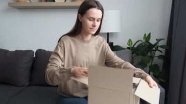 Happy cute young woman sitting on cozy sofa, unpacking box with wished item, feeling satisfied with online shopping or fast delivery service. Smiling beautiful girl unboxing cardboard parcel at home