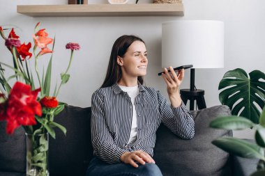 Attractive young woman listening pleasant news in audio message, recording response using cellphone application. Happy female communicating distantly by voicemail sit on couch near flowers at home
