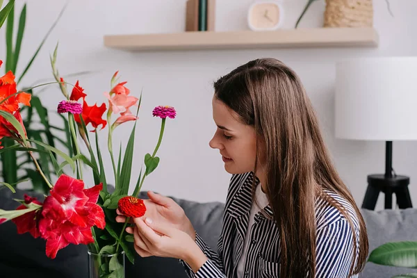 Cute woman enjoys smell of fresh flowers in living room. Happy adult housewife lady doing household activity pouring flowers growing taking care of houseplant cultivation at home. Gardening concept