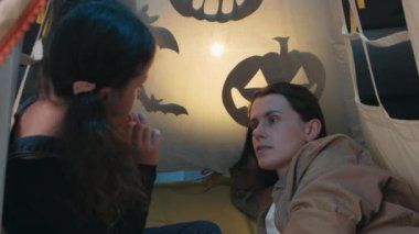 Close up of small daughter and young mother telling spooky stories using shadow play and paper bats and pumpkins. Mom and kid girl have fun, sitting on comfy tent at home. Puppet theater of shadows