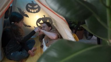 Cute fun little school daughter sitting in cozy tent with shadow bats and spooky pumpkins at home telling stories to loving young mother during Halloween celebration, family spending time together