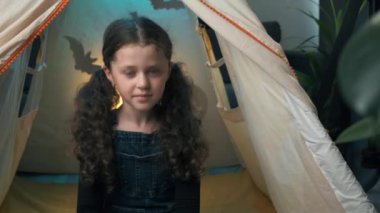 Close up of little child girl blogger looking at camera talking video calling on chatting sitting in dark tent decorated big shadows bats and spooky pumpkins. Blogger and Happy Halloween concept