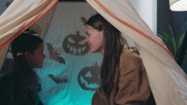 Fun young mother and cute little daughter sitting dark in tent, tell scary stories on background of spooky pumpkins, enjoying bonding cuddling hugging spending time at home. Happy Halloween concept