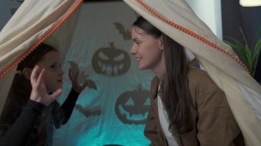 Close up of fun young pretty mother with cute little daughter kid sitting in dark tent at home with funny shadows of bats and big paper spooky pumpkins, have fun together. Happy Halloween concept
