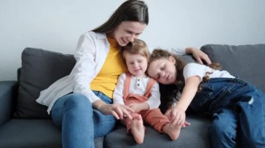 Cheerful young mom and small childs having fun. Playful caucasian mum hugging funny cute little daughters enjoying playing lifestyle game, laughing sitting on comfy grey couch in living room at home
