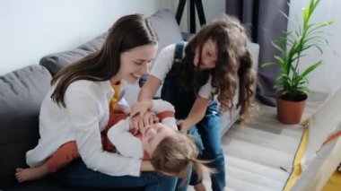 Cheerful young mother and small childs having fun. Happy playful mum hugging funny cute little daughters enjoying playing lifestyle game, laughing sitting on comfortable sofa in living room at home