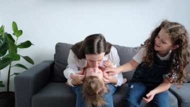 Playful young mother playing with two cute little daughters, mom enjoy time on weekend with cheery kids girls tickles laughing together seated on cozy couch at home, activities with child concept