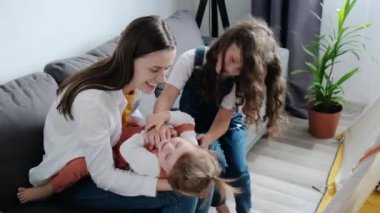 Cheerful young mother and small childs having fun. Happy playful mum hugging funny cute little daughters enjoying playing lifestyle game, laughing sitting on comfortable sofa in living room at home