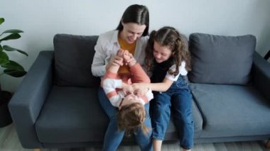 Happy young mom playing with two cute little daughters, mother enjoy time on weekend with cherry kids girls tickles laughing together seated on cozy sofa in living room, activities with child concept