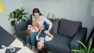 Happy loving young mother piggyback sitting on cozy couch with playful two cute little daughters, family resting at home on comfy sofa, love bonding, caring concept. Sweet tender moment concept