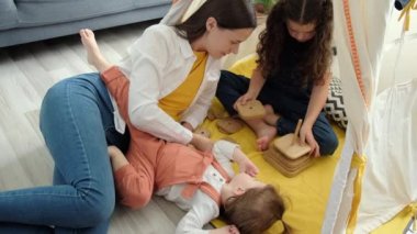 Smiling young mother helping cute little childs daughters playing toys together, beautiful caring mom and small pretty kids girls having fun laugh building constructor tower of wooden blocks at home