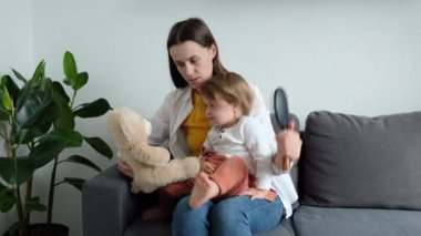 Caring young mother combs daughter hair. Loving mom does hairstyle of cute little girl child, kid holding fluffy bear sitting on comfy sofa in living room at home. Child Care and motherhood concept