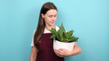 Cheerful young woman in uniform holding plant, posing isolated over blue color background wall in studio. Happy caucasian girl 20s old years posing with flowerpot on backdrop. Gardening concept