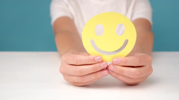 テーブルで黄色の幸せな笑顔の顔を保持している女性の手の閉じる 良いフィードバック評価と肯定的な顧客レビュー 精神健康評価 コピースペースと青のスタジオの背景の壁にポーズ — ストック動画
