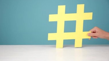 Kadın ellerine yakın büyük sarı hashtag sembolü beyaz masada, esrar işareti ünlü medya içeriği, sosyal medya pazarlaması ve blog promosyonu, mavi arkaplan duvarının üzerinde poz