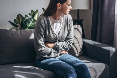 Unhappy young female 20s old years sitting on sofa in living room at home, suffering from pain of abdominal cramps, breathing heavily, clutching holding stomach, gastritis, abdominal, climax concept clipart