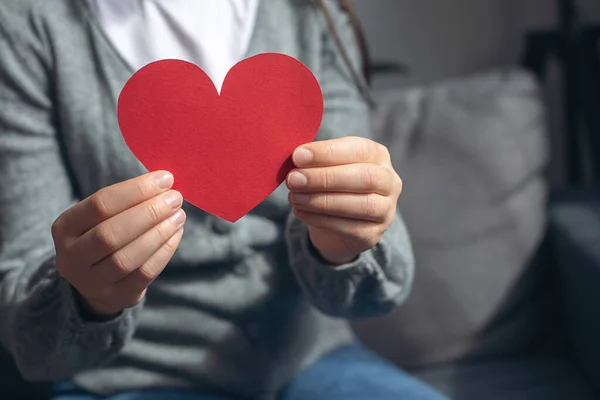 World heart day, world health day, CSR responsibility, adoption foster care home, all lives matter, no to racism concept. Close-up of young female holding small red paper heart sitting on comfy couch