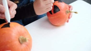 Tatil hazırlığı ve el yapımı parti dekorasyonu. Cadılar Bayramı aile içi aktiviteler, tanınmayan anne-kız balkabağı boyama ve beyaz masaya ürkütücü yüzler çizme. Korkunç bir yüz çiziyor.