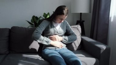 Mutsuz, genç, beyaz bir kadın, gri kanepesinde oturuyor, karın kramplarından muzdarip, derin nefes alıyor, karnına tutunuyor, mide iltihabı, karın ağrısı, orgazm, yüzü acıdan bozulmuş.
