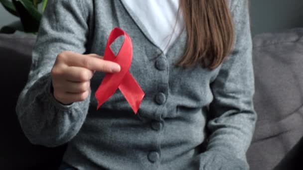 若い女性の閉じる小さな赤いリボン乳がんのシンボルを保持ソファの上に座っている病気の患者をサポートし 女性ボランティアエイズHiv疾患の認識 医学の概念を維持 — ストック動画