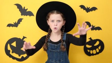 Şapkalı küçük bir kızın portresi, el kaldırıyor ve kameraya bakıyor. Stüdyonun sarı arka plan duvarında izole bir şekilde poz veriyor. Siyah kağıt balkabağı ve yarasalar. Mutlu Cadılar Bayramı konsepti