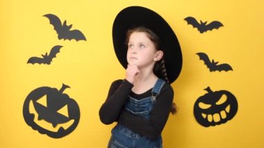 Düşünceli küçük bir kız çocuğu karar vermeyi düşünür, bir şeyler düşünür, sarı stüdyo arka planında izole edilmiş bir şekilde poz verir reklam için fotokopi alanı bırakır. Mutlu Cadılar Bayramı ve garip bir şey