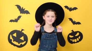 Siyah şapkalı, dans eden, gülümseyen, kameraya bakan, sarı arka planda poz veren, komik balkabağı ve yarasalarla süslenmiş küçük bir kız çocuğu. Parti tatil konsepti