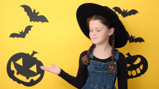 正面的小女孩戴着帽子 用食指指尖在复制空间上展示促销内容 在装饰过的黑色南瓜和蝙蝠工作室的黄色背景上摆出孤立的姿势 万圣节快乐的概念 — 图库视频影像