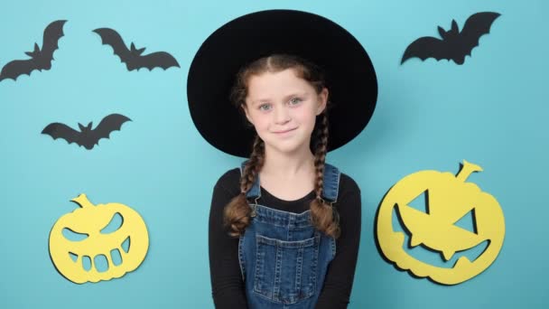 照片中 穿着黑色帽子的可爱小女孩面带微笑 看着相机 在装饰着巨大纸制南瓜和小蝙蝠的工作室里 在蓝色的背景墙上摆出孤立的姿势 万圣节快乐的概念 — 图库视频影像