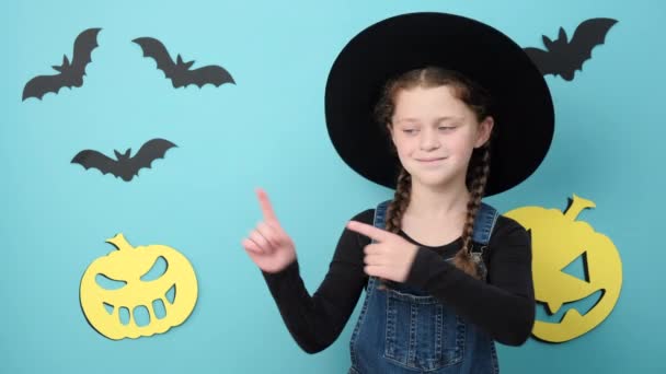 戴着帽子的快乐小女孩的画像 用食指指尖在复制空间上展示促销内容 在工作室装饰南瓜和蝙蝠的蓝色背景下显得孤立无援 秋天和万圣节的概念 — 图库视频影像
