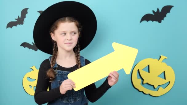 快乐可爱的小女孩戴着黑色的帽子 带着黄色箭头出现在旁边 微笑着看着相机 在蓝色的工作室背景上摆出孤立的姿势 并为促销内容提供复制空间 万圣节的概念 — 图库视频影像
