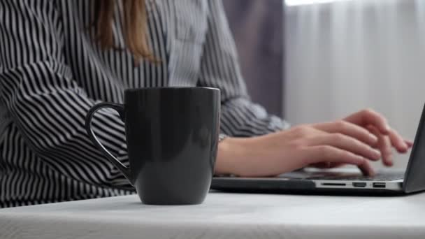 ノートパソコンをタイプする女性の手を閉じて 窓の近くの机の上に熱いコーヒーカップを持っています 女性のフリーランサーはコンピューターで働いてる ソーシャルネットワーク 小規模ビジネス スタートアップのコンセプト 選択的焦点 — ストック動画