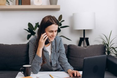 Bilgisayarda çalışan gülümseyen genç bir kadın cep telefonu danışmanı müşterisi ya da internet müşterisi olarak rahat bir koltukta oturuyor. Mutlu iş kadınının ortağıyla akıllı telefon görüşmesi var..