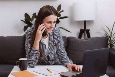 Kanepede dizüstü bilgisayar kullanarak oturan ve akıllı telefondan konuşan mutlu genç bayan. Gülümseyen kız arkadaşlarıyla sohbet ediyor, bilgisayar teknolojisiyle evde çalışıyor rahat bir kanepede dinleniyor. Başlangıç konsepti