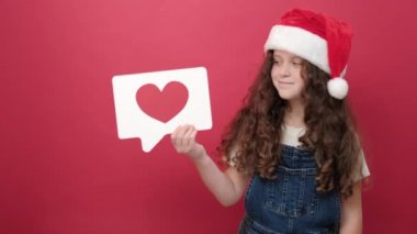 Noel Baba şapkası takan küçük bir kızın portresi sosyal medyada ikon gibi ayakta duruyor, kırmızı arka planda izole edilmiş içeriği takip etmeyi ve sevmeyi öneriyor. Mutlu yıllar 2023 tatil konsepti