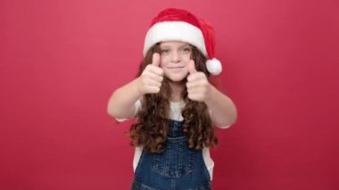 Gülümseyen küçük kız portresi başparmak kaldırıyor, Noel Baba şapkası ve beyaz tişört giyiyor, stüdyo odasındaki kırmızı renkli arka plan duvarında fotokopi alanı ile izole edilmiş poz veriyor. Mutlu yıllar tatil konsepti