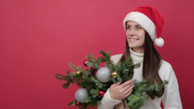 Mutlu genç Noel Baba kadın beyaz süveter giyer ve Noel şapkalı ladin demeti bir yana bakar, stüdyodaki kırmızı arkaplan duvarının üzerine izole edilmiş poz verir. Mutlu Yıllar Kutlama Konsepti