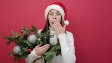 Şok olmuş genç bir kadının portresi beyaz süveter giyer ve Noel Baba şapkası takar ladin dallarının buketi ağızlarını eliyle kapatır, kırmızı stüdyo arka plan duvarında izole bir şekilde poz verir. Mutlu yıllar tatil konsepti