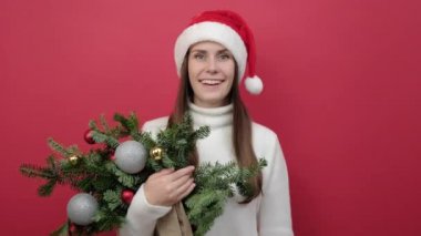 Mutlu 20 'li yaşlarda genç bir kadın Noel Baba şapkası ve süveter buketi giyer ladin dalları buketi parmağıyla sizi motive eder cesaretlendirir, stüdyonun kırmızı arka plan duvarında izole bir şekilde poz verir.