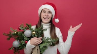 Mutlu yeni yıl kutlamaları. Noel şapkalı mutlu genç Noel Baba kadın elinde bir demet ladin dallarıyla kırmızı renk arka plan stüdyosunda fotokopi alanı ile poz veriyor.