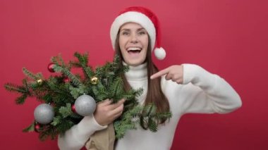 Şaşırmış genç bir kadının portresi beyaz süveter giyer ve Noel Baba şapkası, stüdyodaki kırmızı arkaplan duvarının üzerine izole edilmiş ladin dalları buketinde durur. Mutlu yıllar 2023 tatil konsepti