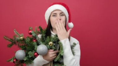 Çekici genç kadın portresi beyaz süveter giyer ve Noel Baba şapkası takar ladin dalları üfler, hava öpücüğü flörtü gönderir, stüdyonun kırmızı arka plan duvarında izole edilmiş poz verir.