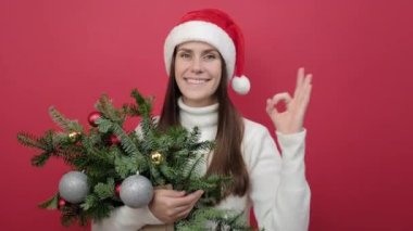 Mutlu 20 'li yaşlardaki genç kadın portresi beyaz süveter giyer ve Noel Baba şapkası demetinde ladin dalları bulunur. Stüdyonun kırmızı arka plan duvarında izole edilmiş şekilde poz verir. Xmas kavramı