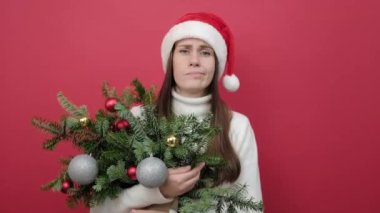 Kafası karışmış beyaz kazaklı beyaz kadın portresi ve elinde ladin dallarından bir buket tutan Noel Baba şapkası kırmızı arka plan stüdyosuna izole edilmiş fotokopi alanı ile yayılmış. Yeni yıl tatili konsepti