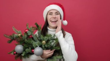 Genç esmer kadının portresi beyaz süveter giyiyor ve elinde ladin demeti tutan Noel Baba şapkası var. Ağız kenarında indirimli satış yapan eller hakkında çığlıklar atıyor. Kırmızı arka plan stüdyosunda izole edilmiş poz veriyor.