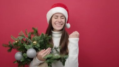 Neşeli 20 'li yaşlarda genç bir kadın süveter giyiyor ve elinde bir buket ladin çiçeği tutan Noel Baba şapkası var. Kazanan jestler yapıyor. Yumruklarını sıkmanı kutluyor.
